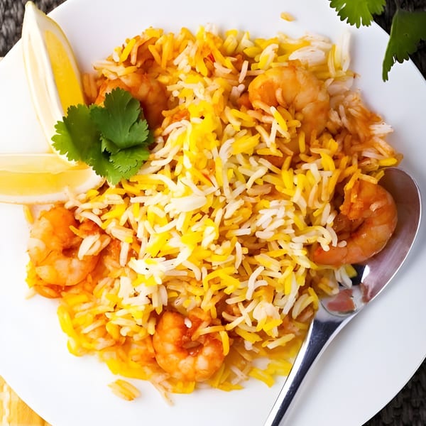 Shrimp biryani with yellow and white rice, garnished with cilantro and lemon wedges on a white plate.