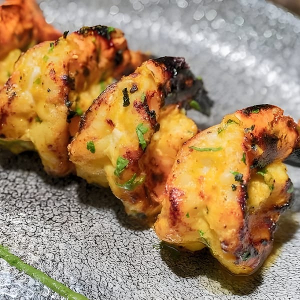 Close-up of grilled Indian shrimp skewers with charred edges and green herbs on a textured plate.