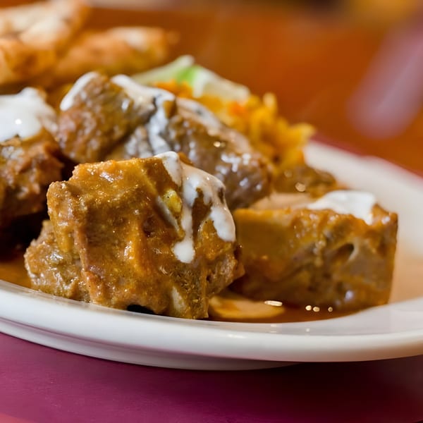 Close-up of Indian curry with meat chunks, drizzled with white sauce, on a white plate.