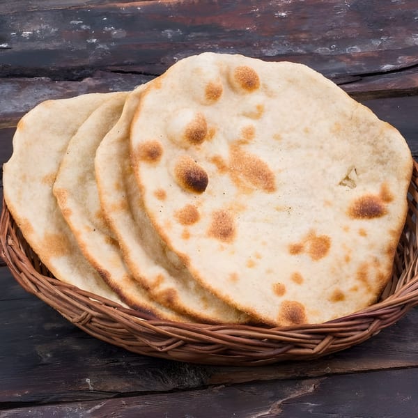 A wicker basket filled with several pieces of freshly baked, golden-brown Indian flatbread on a dark wooden surface.